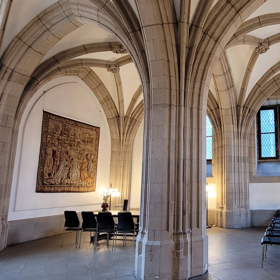 Abgebildet ist ein Trauzimmer mit historischen hohen Rundbögen sowie einem Trautisch mit Stühlen an der linken Seite des Bilds. Weiterhin sind Stühle für Gäste zu sehen, die an der rechten Seite mit Blick zum Hochzeitspaar angeordnet sind.